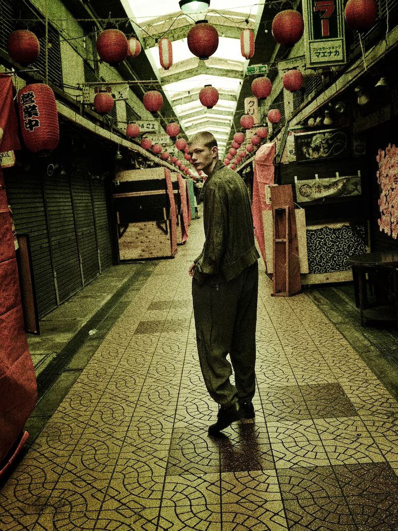 Man in loose green outfit glances back while walking down an outdoor covered market aisle adorned with red lanterns in a green color grade.