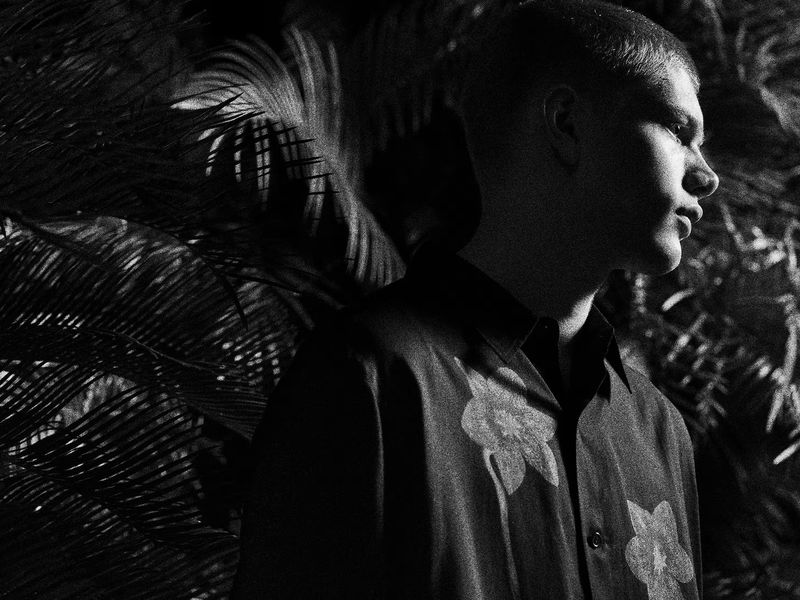 Monochrome portrait of a young adult male in floral shirt, profile view, against dark backdrop of ferns. Black and white photography.