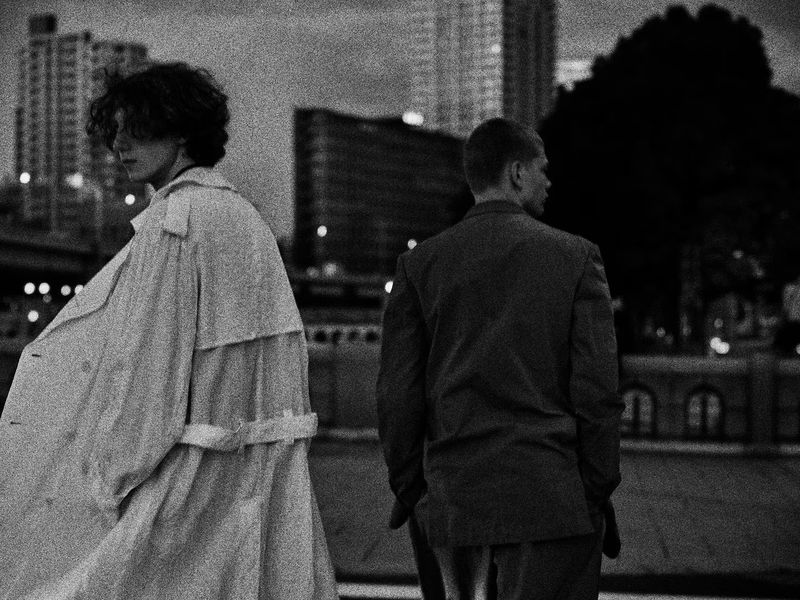 Two models outdoors at night wearing oversized coats against an urban city street and building backdrop in a grainy black and white shot.