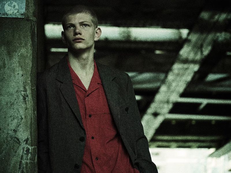 Portrait of a young adult man with a buzzcut, wearing a red shirt and grey jacket, standing in an urban environment with geometric structural elements.