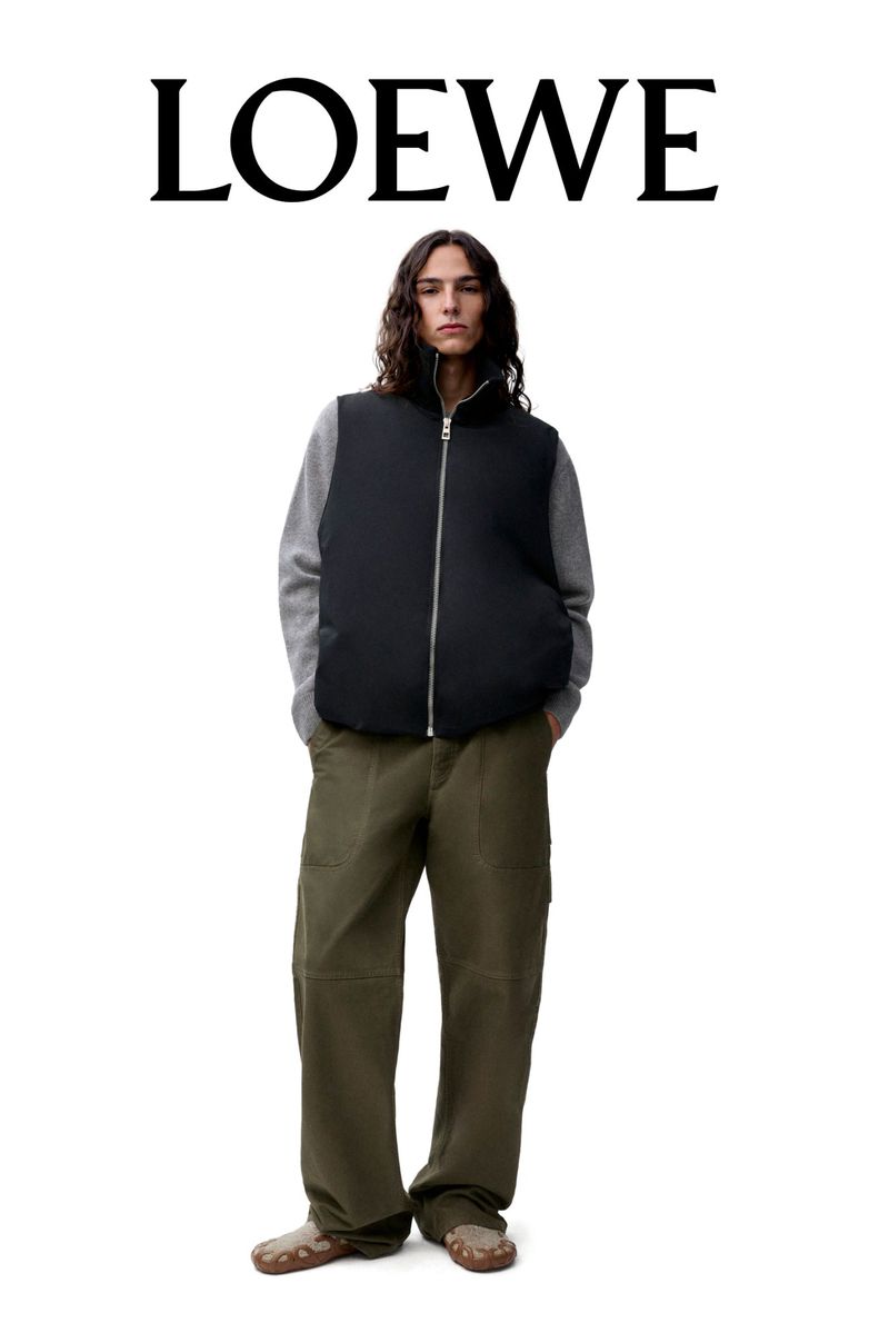 Fashionable adult male with long brown hair posing in a black puffer vest, grey sweater, and olive green pants, with the LOEWE logo.