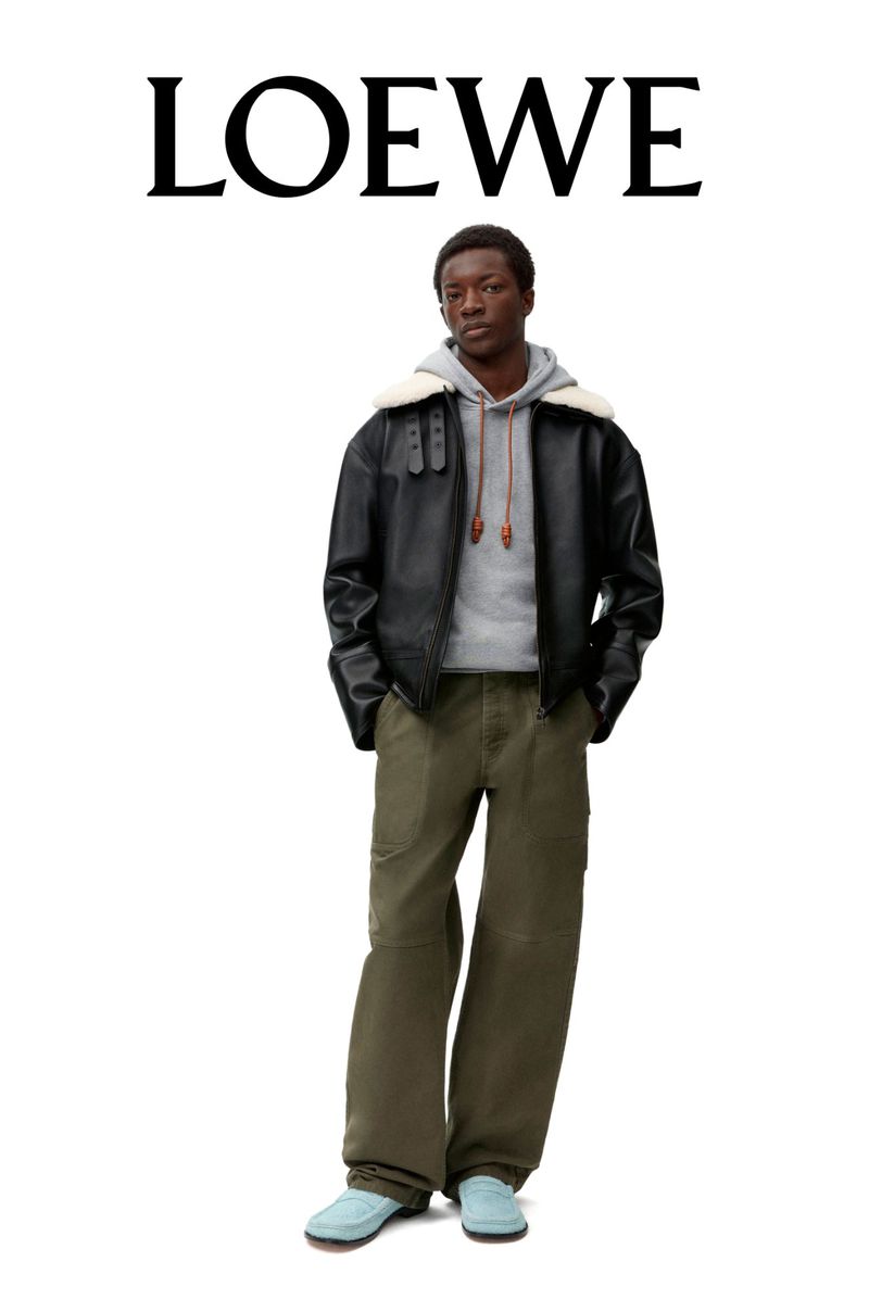 A stylish adult male model wears a black bomber jacket, gray hooded sweatshirt, olive green pants, and light blue shoes in a fashion shot.