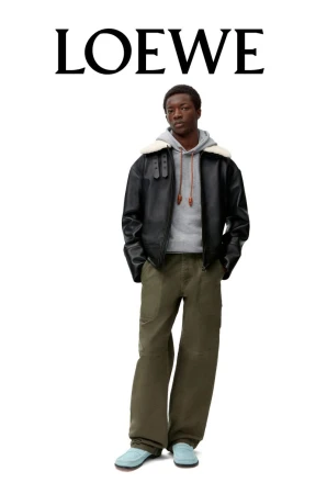 A stylish adult male model wears a black bomber jacket, gray hooded sweatshirt, olive green pants, and light blue shoes in a fashion shot.