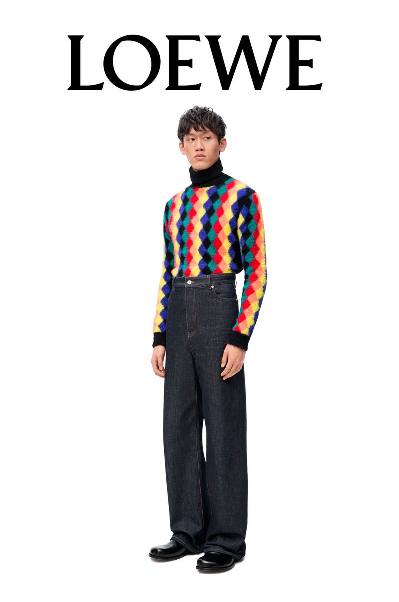 An Asian male model poses in a studio wearing a colorful diamond-patterned sweater, denim pants, and black shoes against a white background.