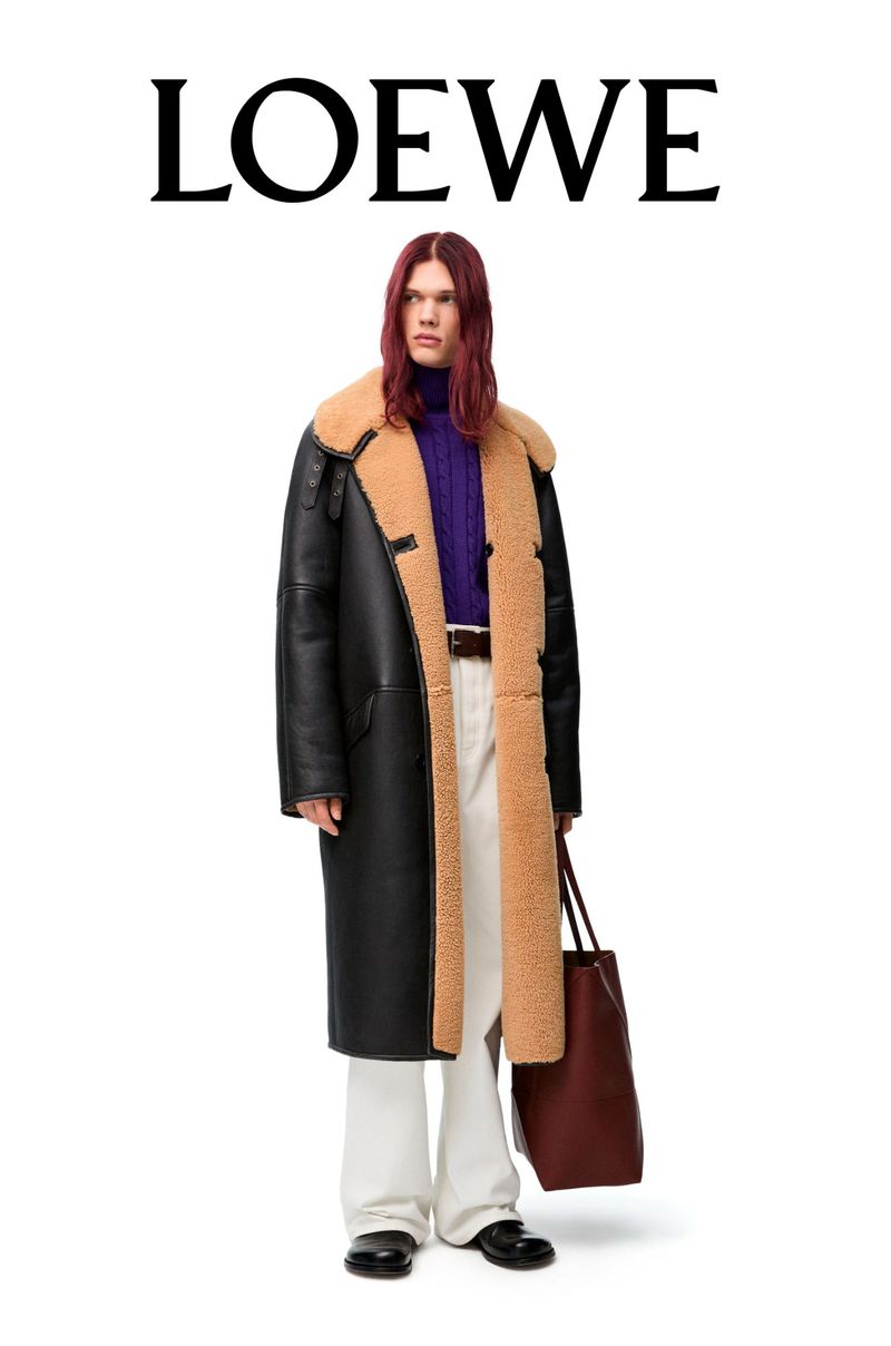 A person with long hair in a purple top and white pants holds a burgundy tote bag and wears a black shearling coat, set against a white backdrop.