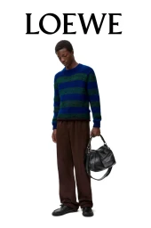 Model standing against a white backdrop wearing a blue striped sweater, brown trousers, and holding a black leather handbag.