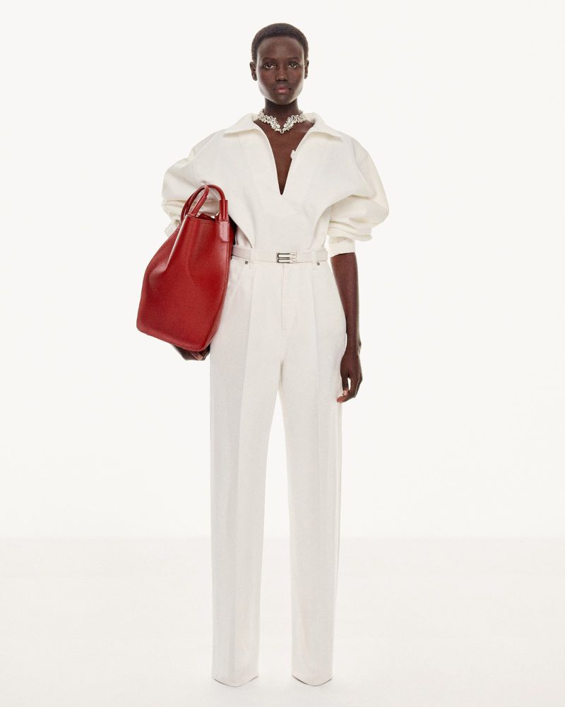 Dark-skinned woman in an off-white jumpsuit with a plunging neckline poses with a red leather handbag in a clean, bright studio.