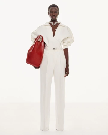 Dark-skinned woman in an off-white jumpsuit with a plunging neckline poses with a red leather handbag in a clean, bright studio.