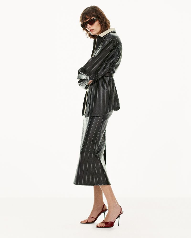 Model in dark sunglasses and a black pinstripe belted jacket with matching skirt against a bright white background.