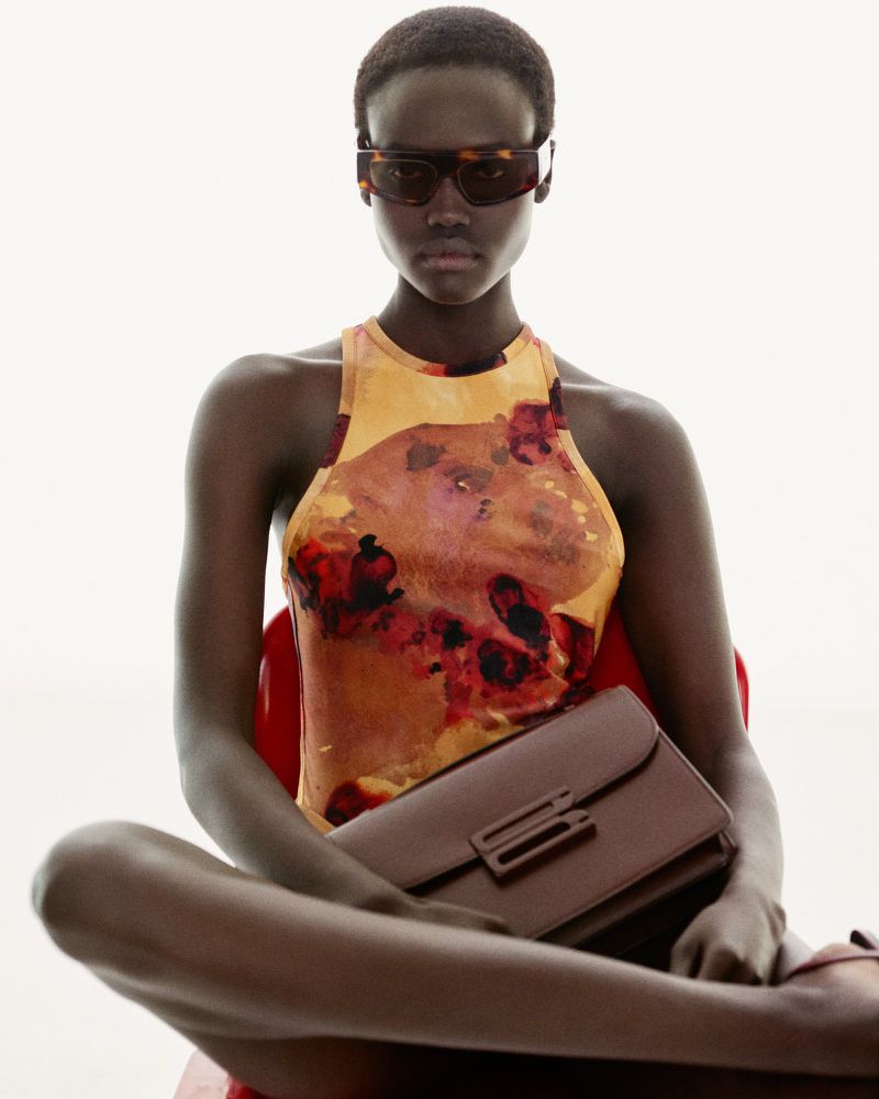 Dark-skinned woman poses seated, wearing brown sunglasses and a floral halter top, holding a dark brown clutch in a studio setting.