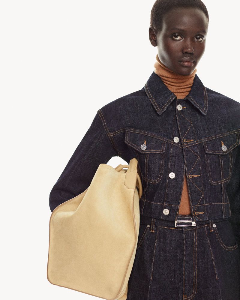 Brunette model wearing a blue denim jacket and skirt carrying a tan leather hobo bag, shot in a bright white studio background.