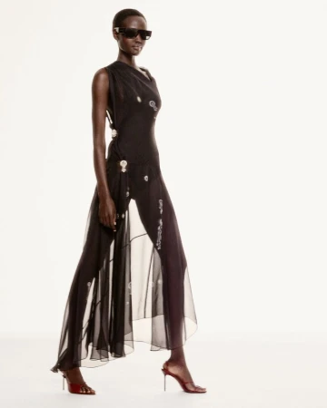 Model wearing dark sunglasses and a sheer black dress against a plain white background