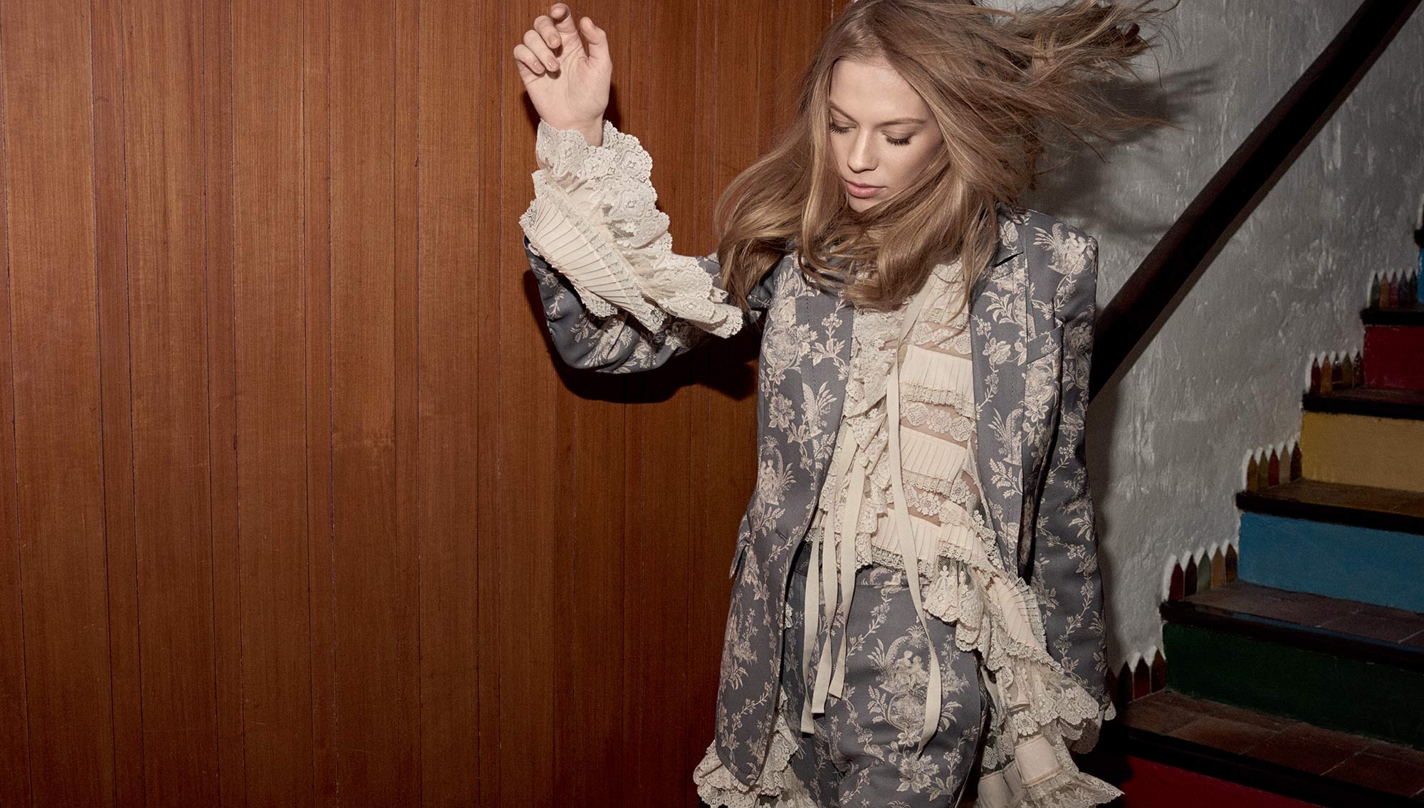 A stylish woman with blonde hair models a floral patterned suit with lace details indoors against a paneled wall and colorful stairs.