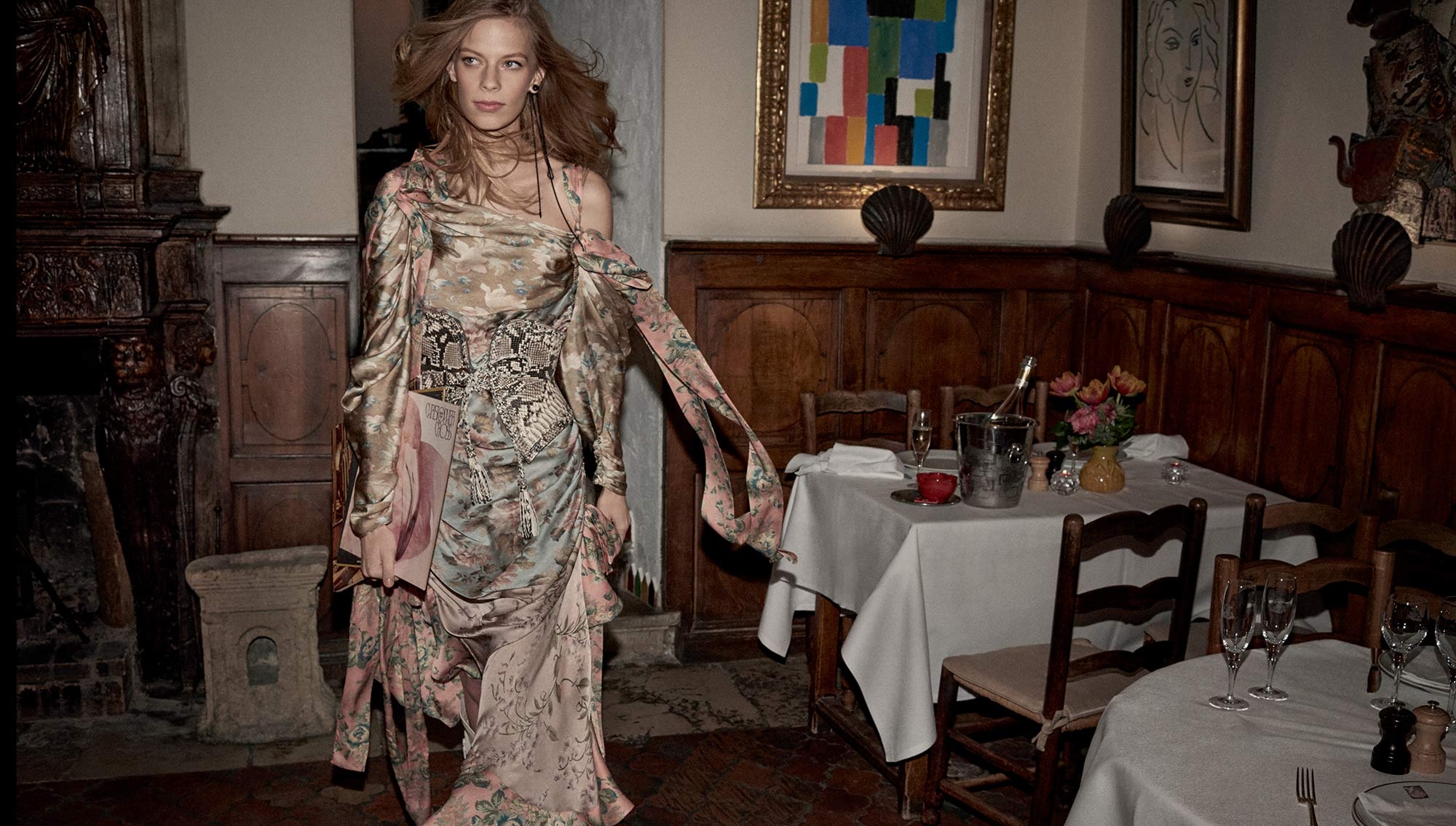 A stylish adult female poses indoors wearing a floral-print gown with an asymmetric neckline and holding a magazine in a dining environment.