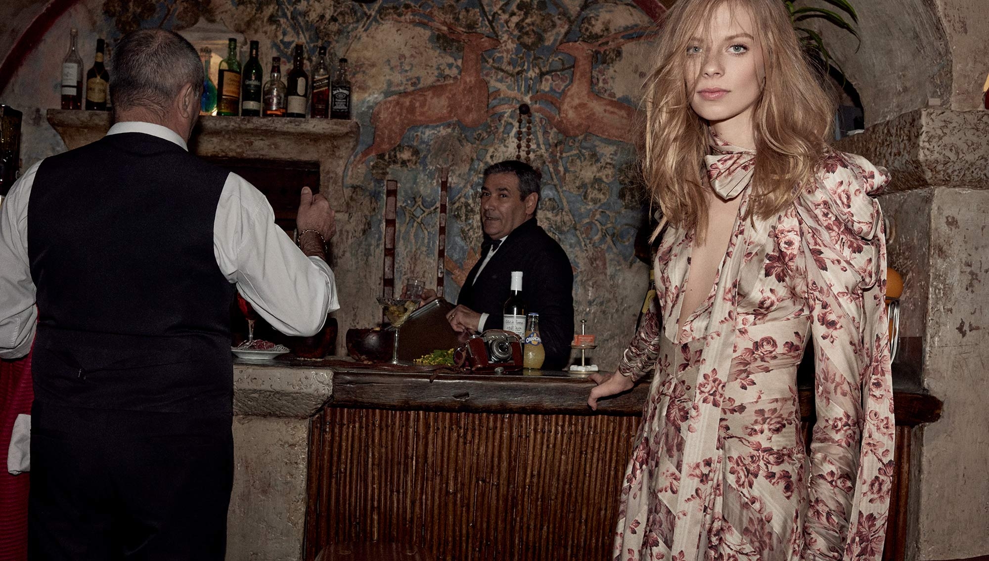 Blond woman in floral dress at rustic bar with two men wearing vests; dimly lit interior with artistic wall paintings.