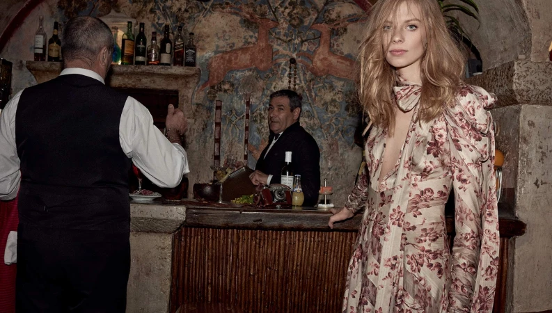 Blond woman in floral dress at rustic bar with two men wearing vests; dimly lit interior with artistic wall paintings.