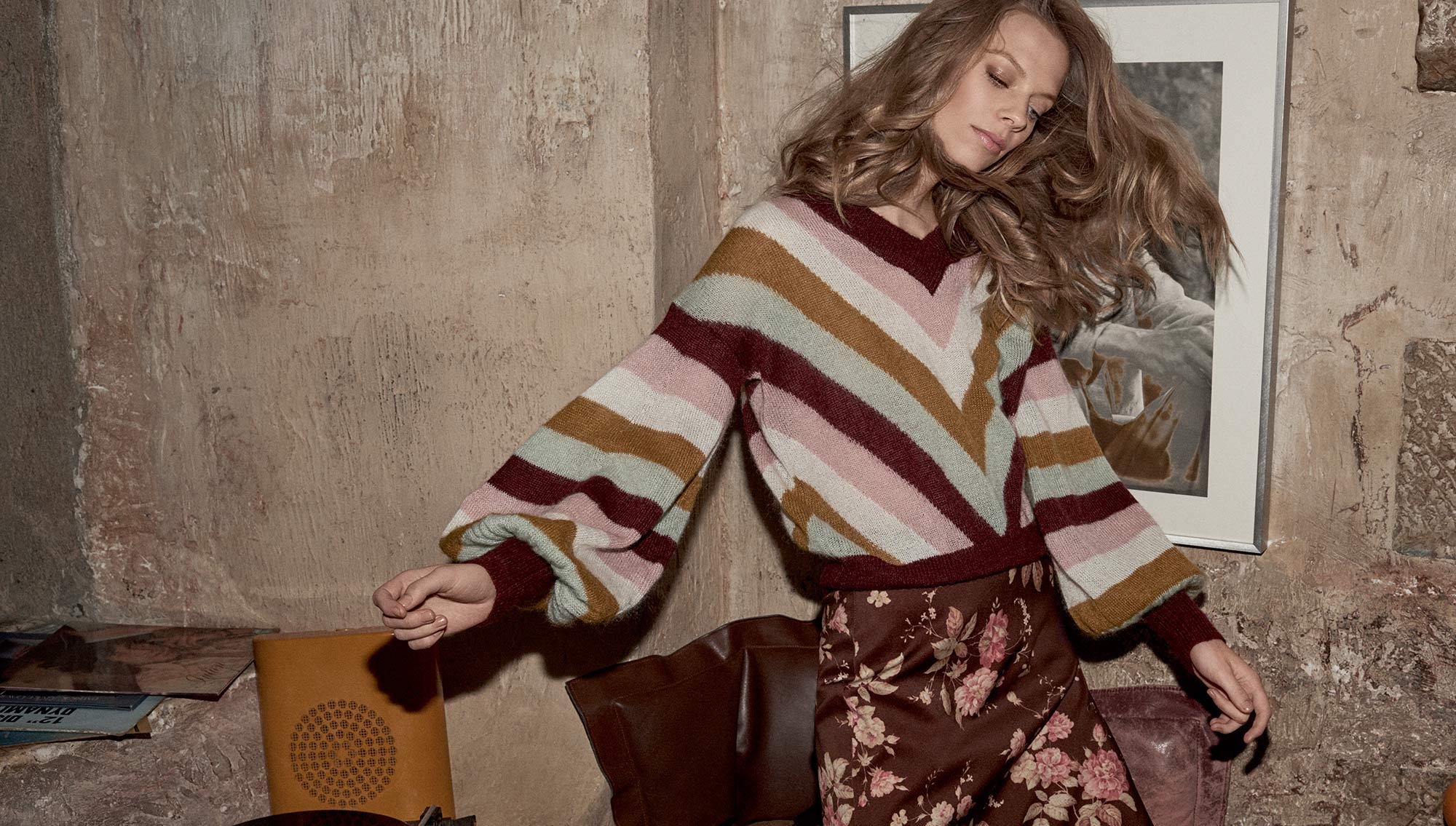 Woman in a striped V-neck sweater and floral skirt posing against a textured stone wall for Zimmermann.