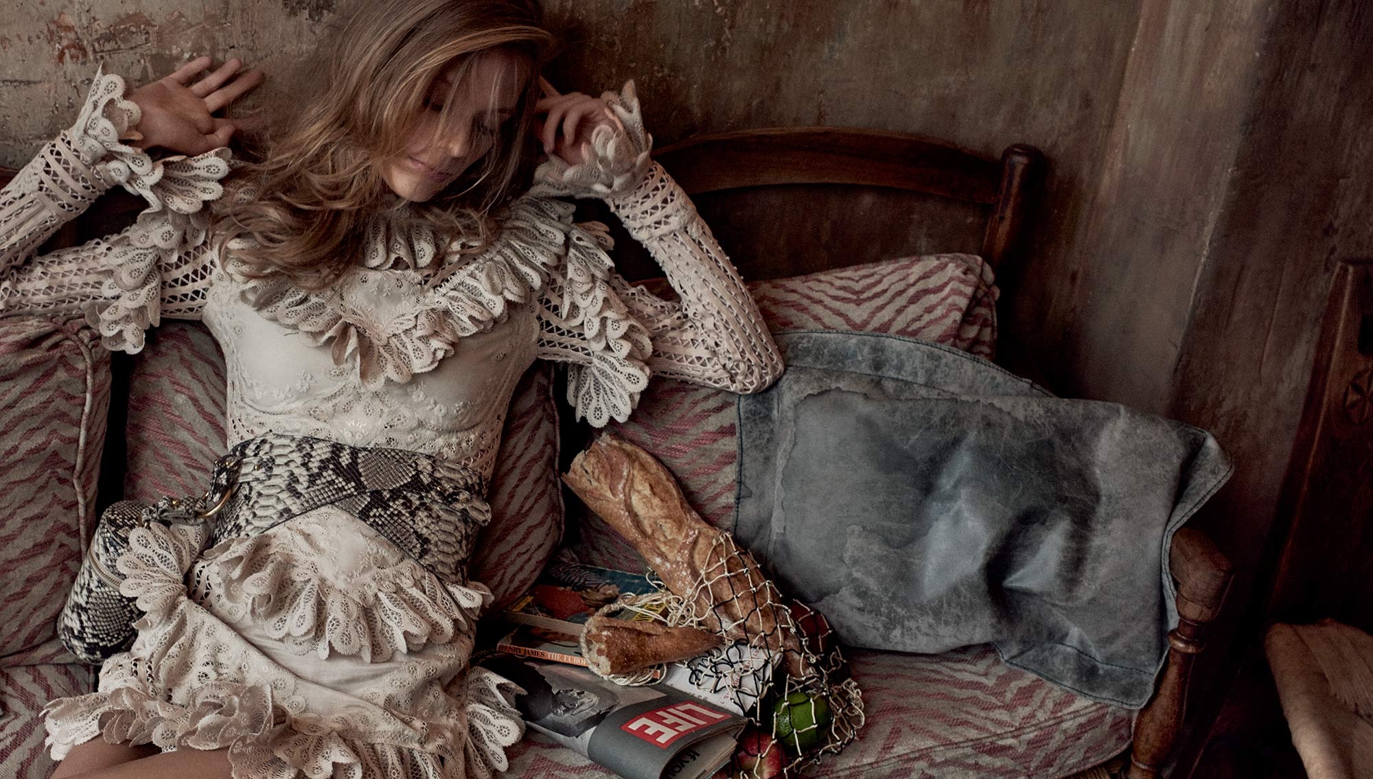 Blonde woman reclines on a vintage patterned couch wearing a white lace dress with a snake-print belt, holding her hand to her face.