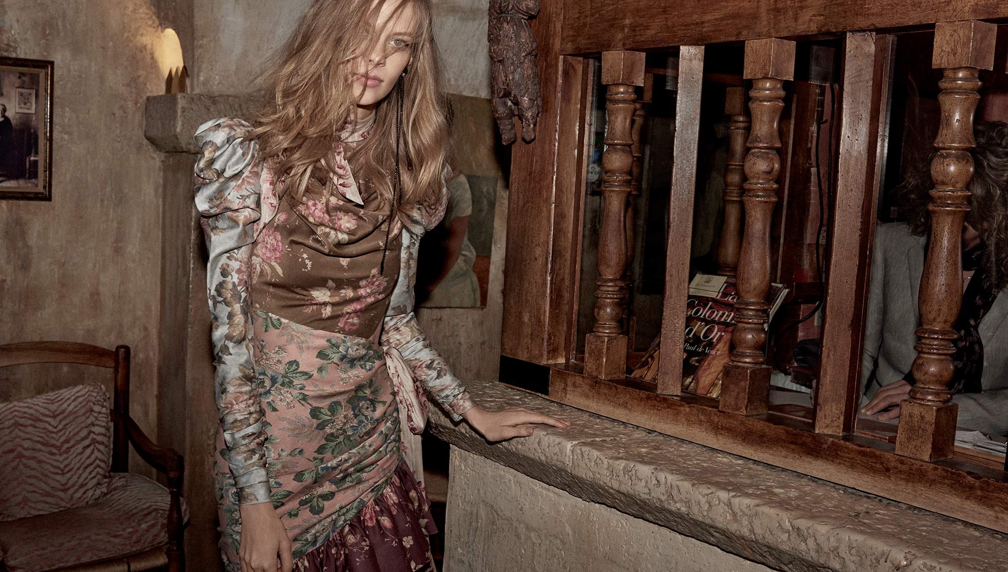 Woman in floral dress posing indoors next to window with wood frame and stone ledge in editorial fashion shot with warm, muted tones.