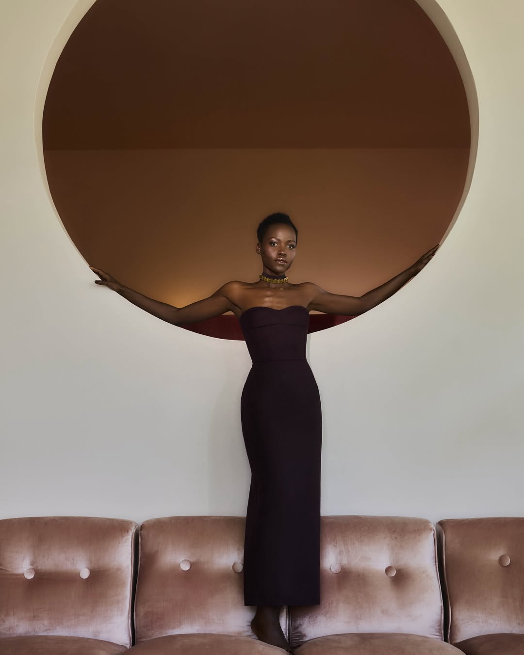Glamorous African woman in a strapless evening gown strikes a pose atop a pink velvet couch in front of a tan circle.
