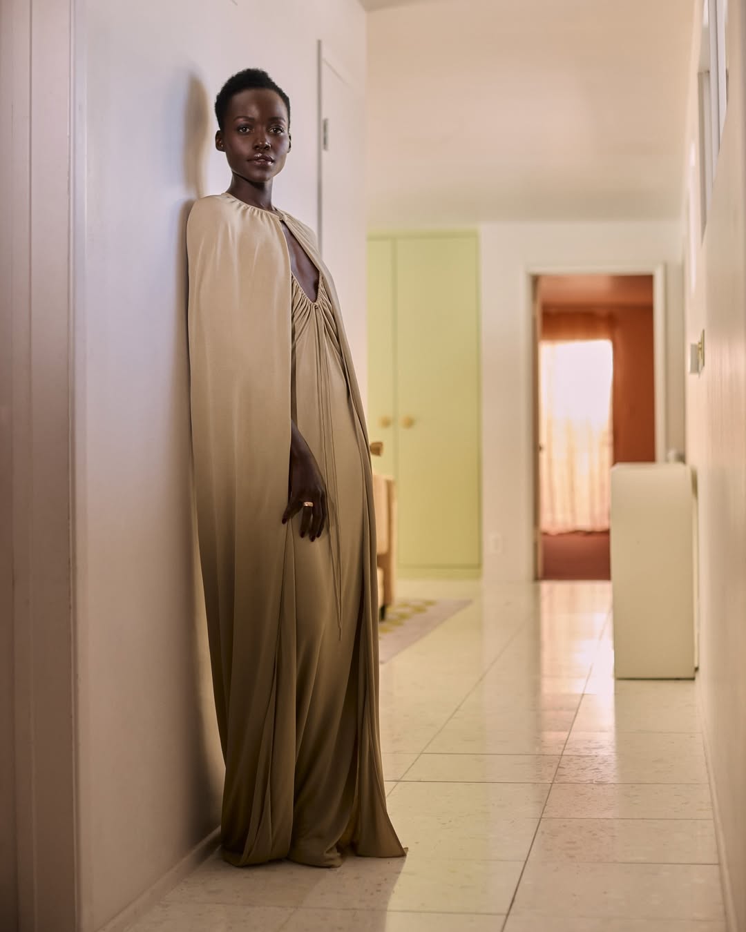 Elegant adult woman in floor-length cape dress stands in interior hallway with neutral tones and natural lighting, fashion photography.