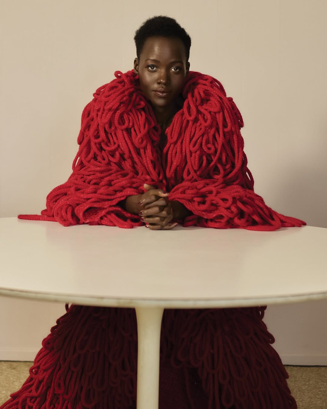 Portrait of Lupita Nyong'o wearing a red yarn garment, seated at a white table against a neutral background in a photo studio.
