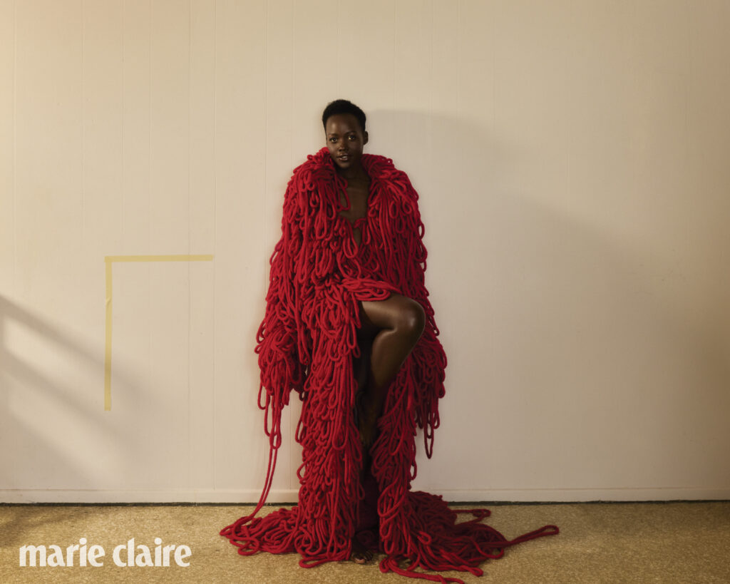 Fashion editorial shot of a woman draped in a red yarn garment against a neutral backdrop, full figure, studio lighting, Marie Claire logo.