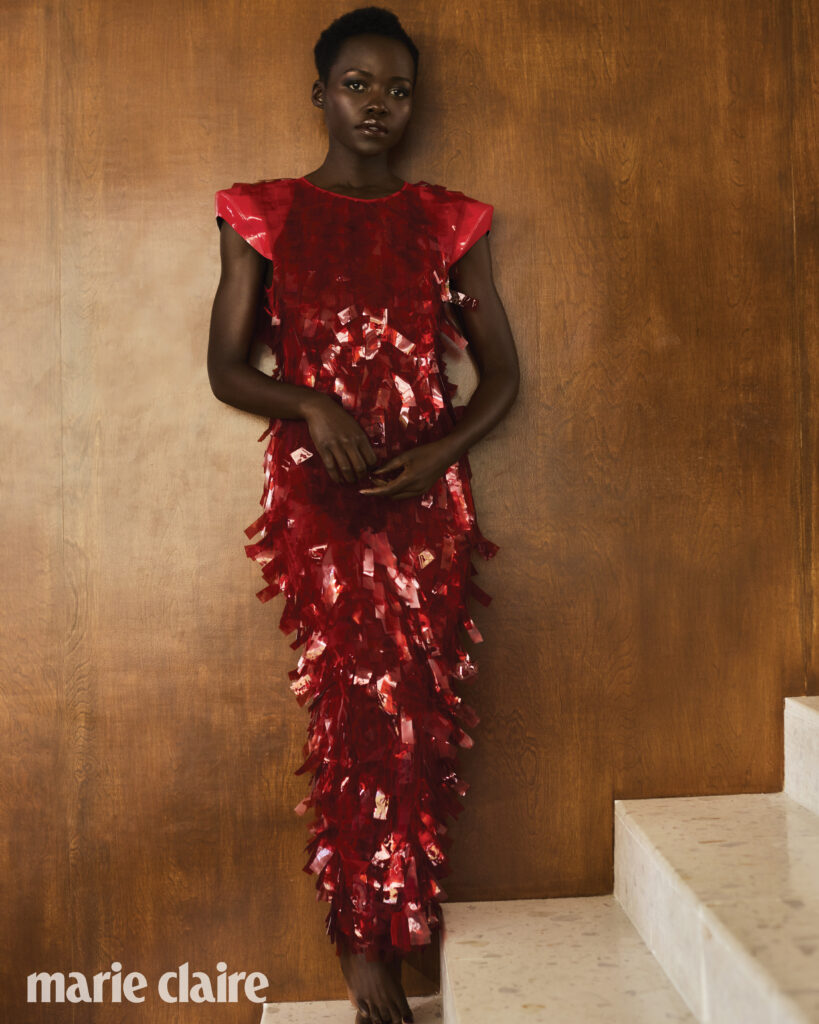 Lupita Nyong'o in a red sequined dress poses against a brown wall and white stairs, showcasing high fashion and editorial style.