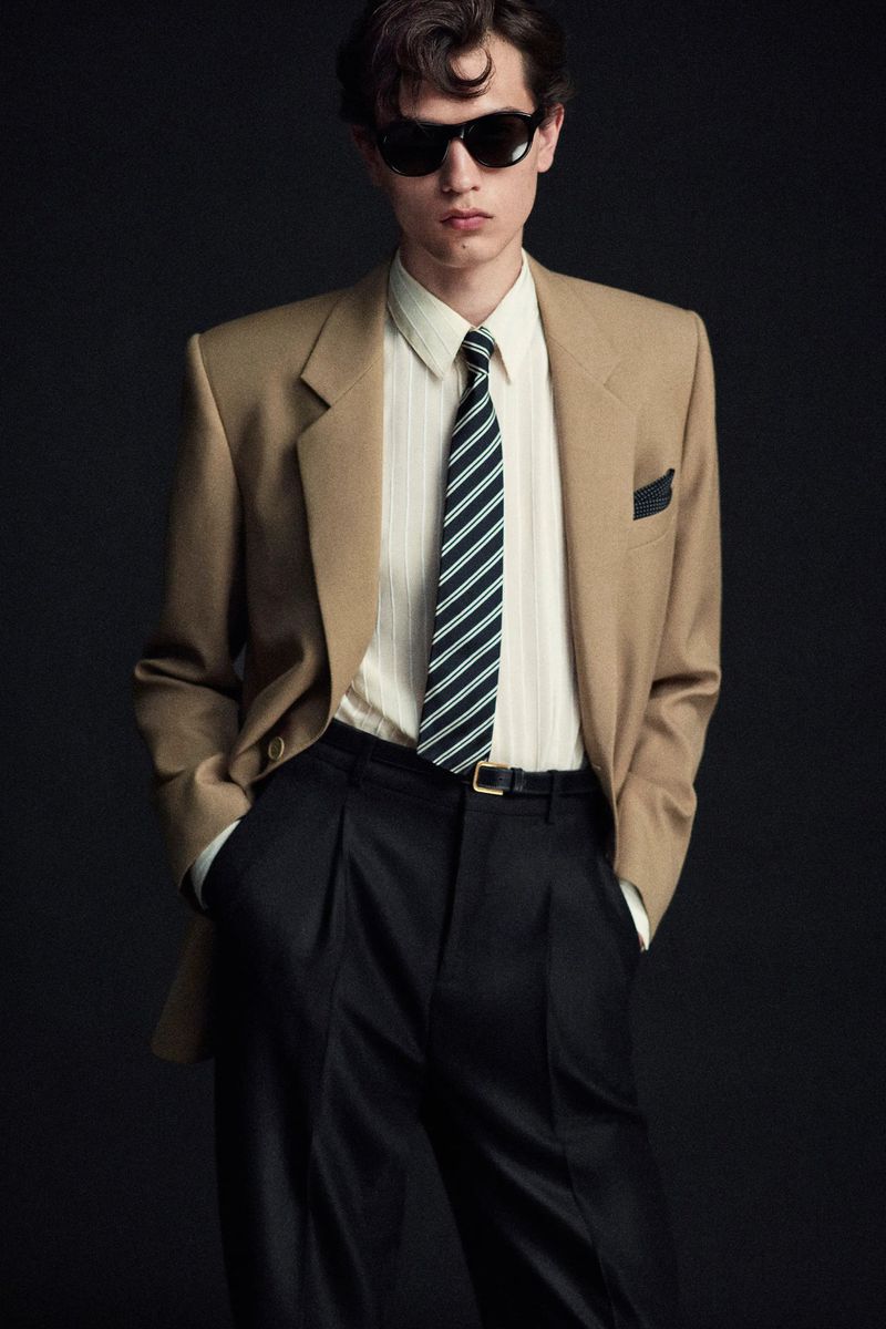 Portrait of a young adult male fashion model in a beige blazer, striped tie, and black sunglasses against a dark backdrop.