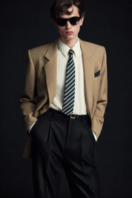 Portrait of a young adult male fashion model in a beige blazer, striped tie, and black sunglasses against a dark backdrop.