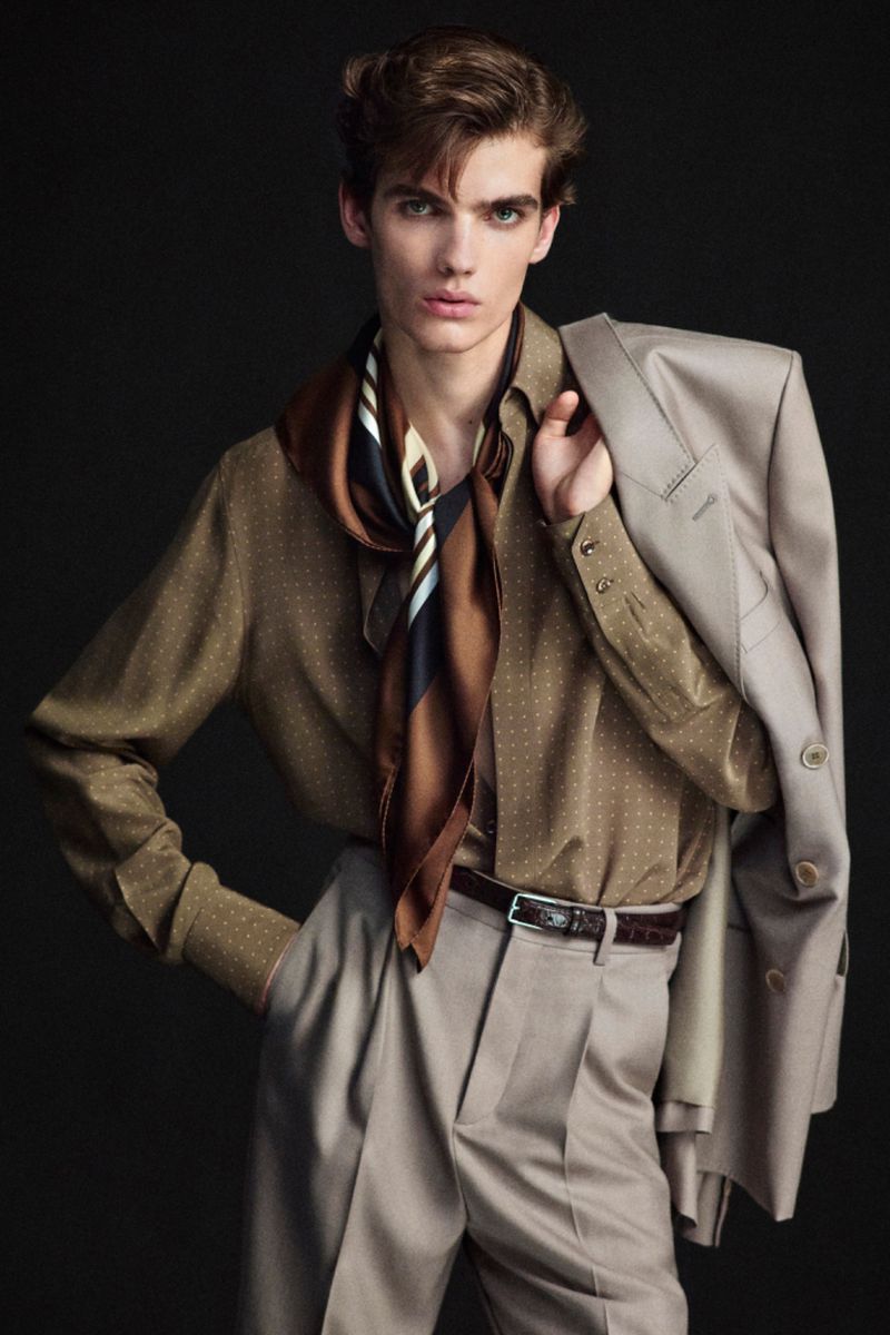 Portrait of a young adult man wearing a tan suit and silk scarf, posing against a dark backdrop in a fashion editorial style.