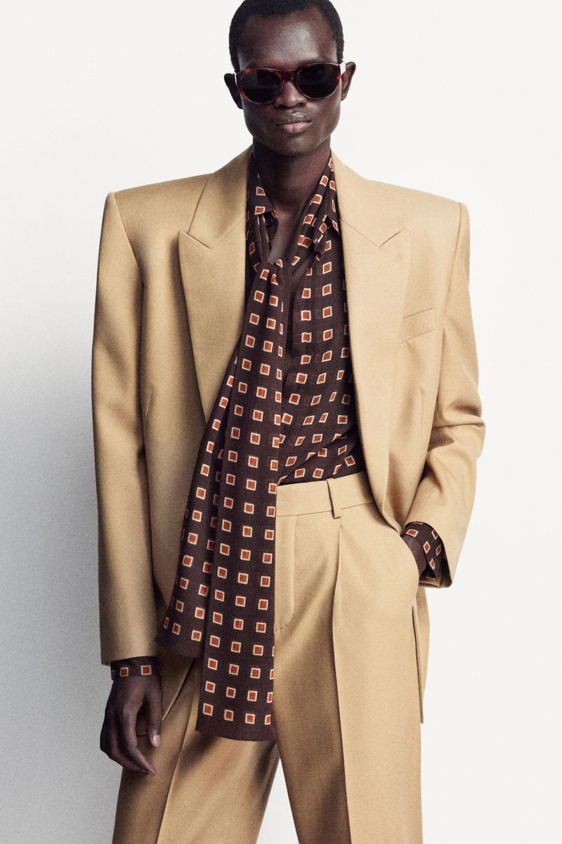 A tan-skinned male model poses in a tan suit with a patterned brown shirt and sunglasses against a white backdrop, fashion photography.
