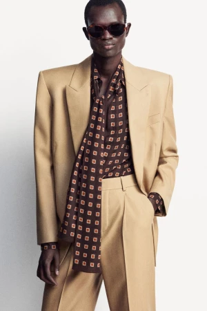A tan-skinned male model poses in a tan suit with a patterned brown shirt and sunglasses against a white backdrop, fashion photography.