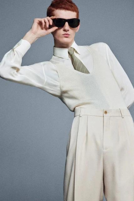 Man with red hair poses in a beige suit vest, long sleeve shirt, tan trousers, and dark sunglasses against a grey backdrop. 