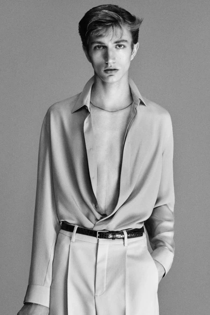 Portrait of a young adult male fashion model in a draped shirt and trousers, posing indoors against a neutral gray background in black and white.