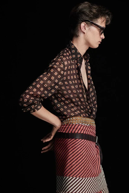 Man posing in a brown patterned sheer shirt, patterned skirt, and sunglasses against a black background in a studio fashion shot.