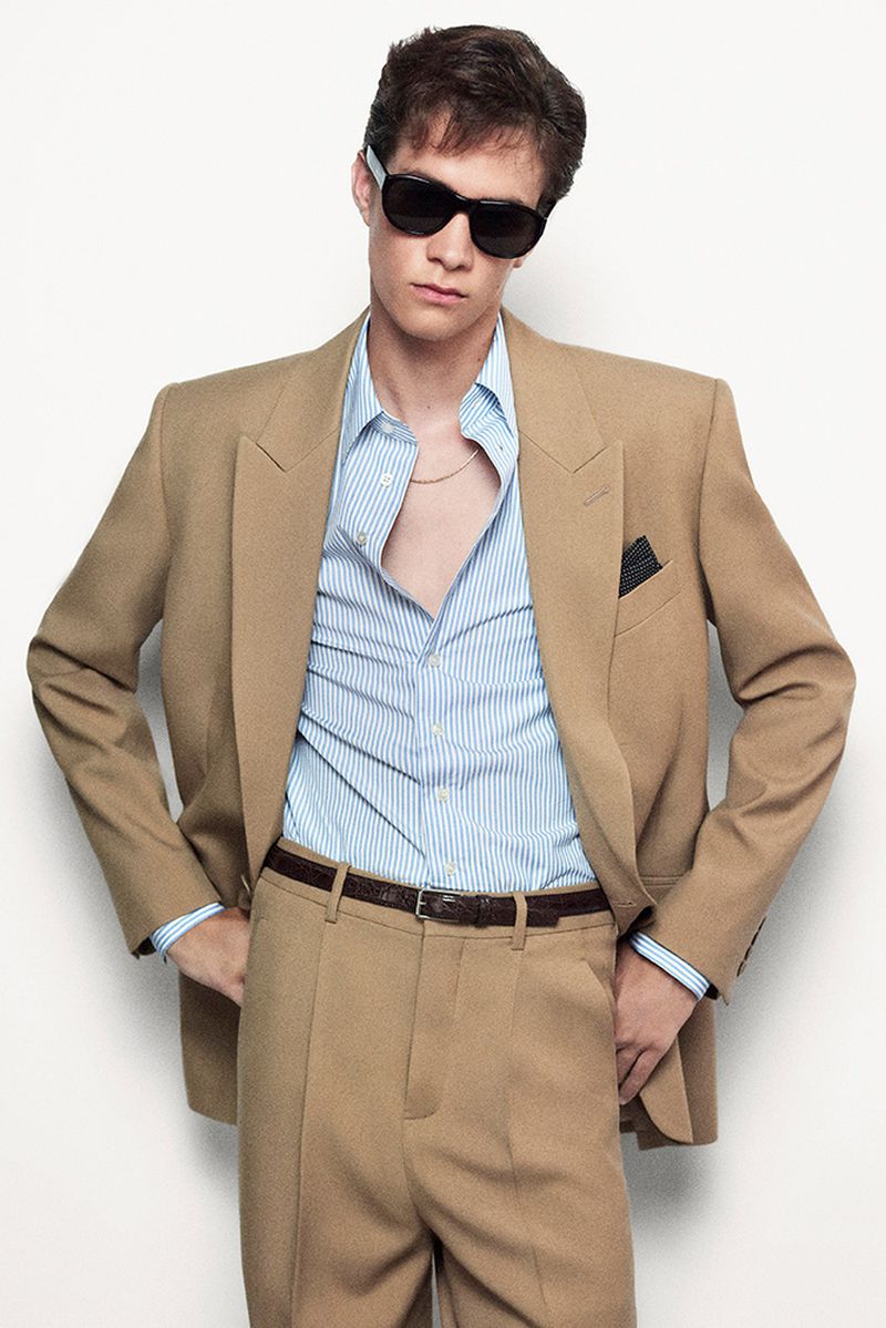 Model wearing dark sunglasses, a tan suit jacket, blue striped shirt, and trousers against a white seamless background.