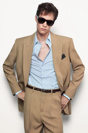 Model wearing dark sunglasses, a tan suit jacket, blue striped shirt, and trousers against a white seamless background.