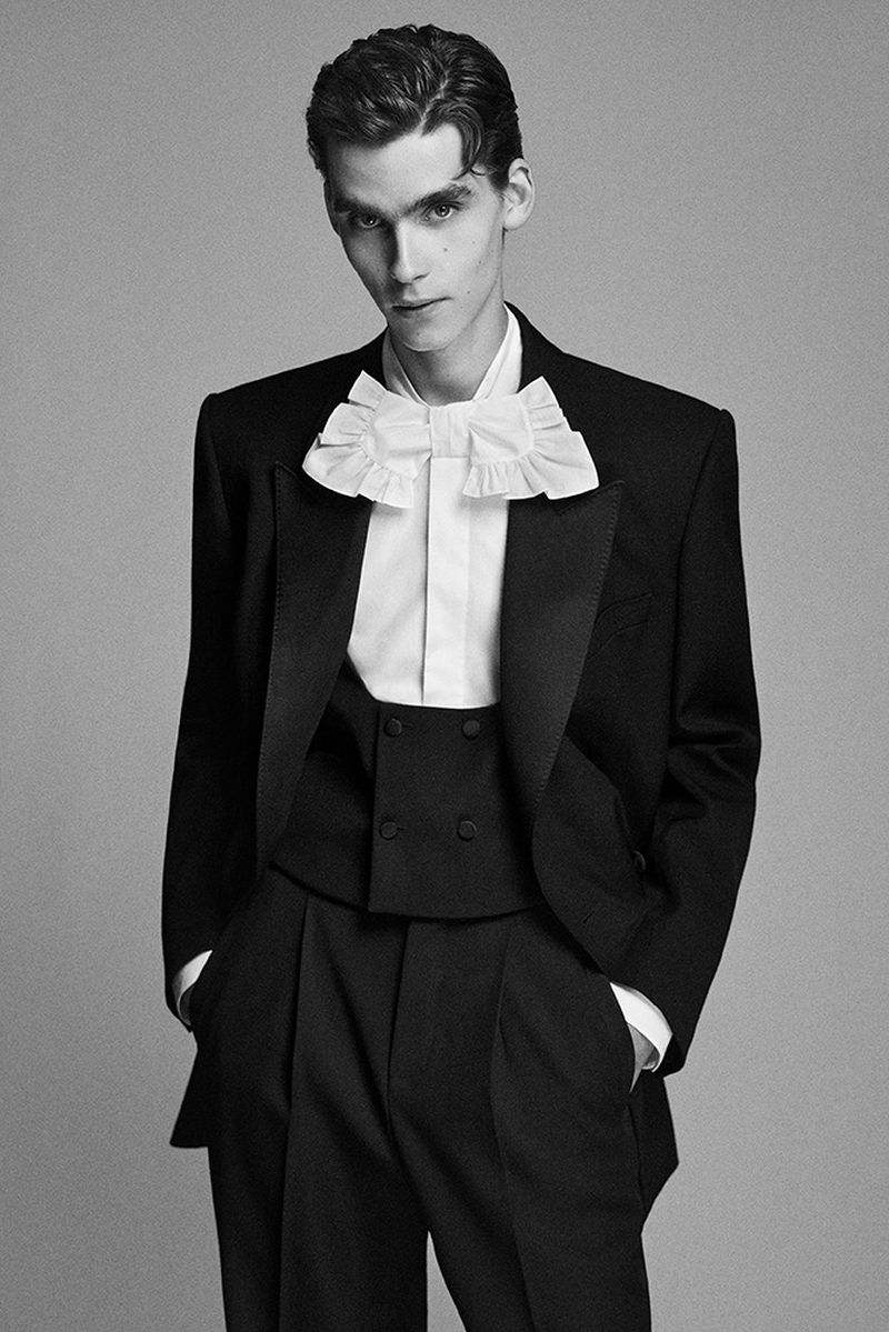Elegant adult male in a black tuxedo with a ruffled white shirt, posing in a studio with a neutral background, portraying a formal look.