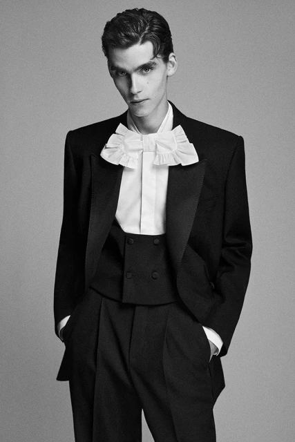 Elegant adult male in a black tuxedo with a ruffled white shirt, posing in a studio with a neutral background, portraying a formal look.