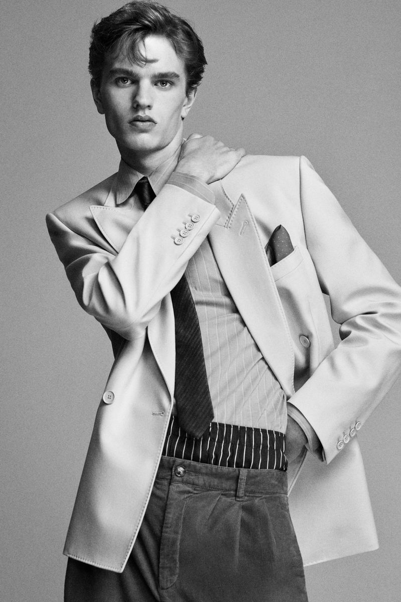 Black and white portrait of a stylish young man wearing a suit jacket, shirt, and tie, posing in front of a neutral background.