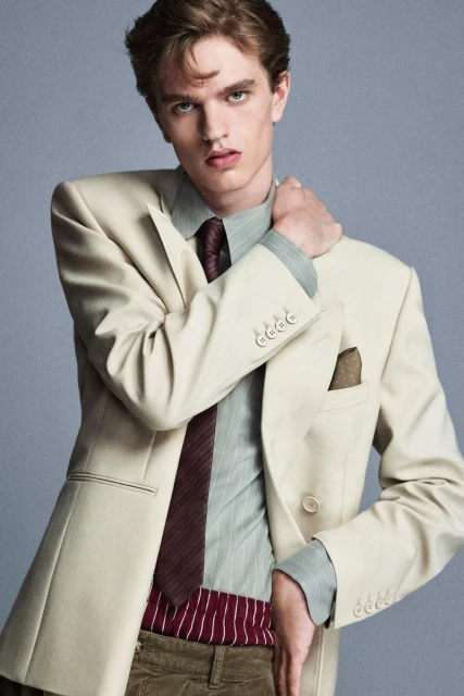 Adult male model poses in a cream-colored double-breasted blazer, striped shirt, and corduroy pants against a light gray backdrop.