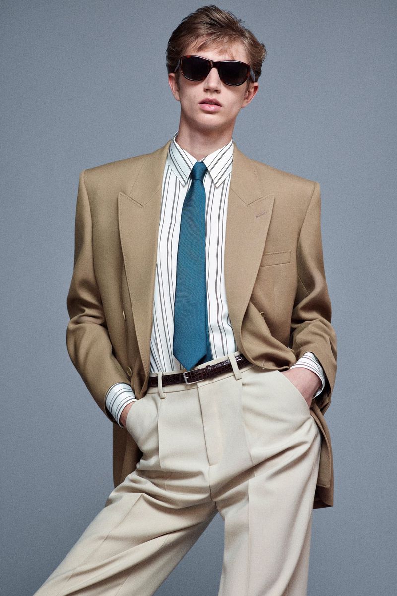 Model wearing dark sunglasses, a beige suit jacket, striped shirt, blue tie, and light trousers against grey.