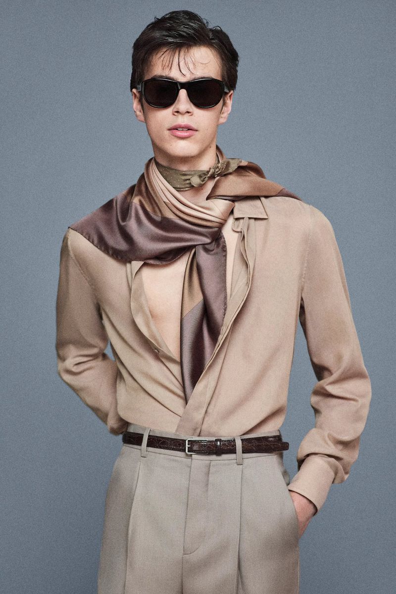 Stylish young man poses in a beige button-down shirt, trousers, and a patterned scarf, accented by sunglasses in a neutral studio setting.