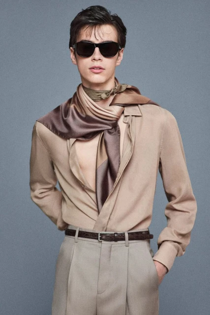 Stylish young man poses in a beige button-down shirt, trousers, and a patterned scarf, accented by sunglasses in a neutral studio setting.