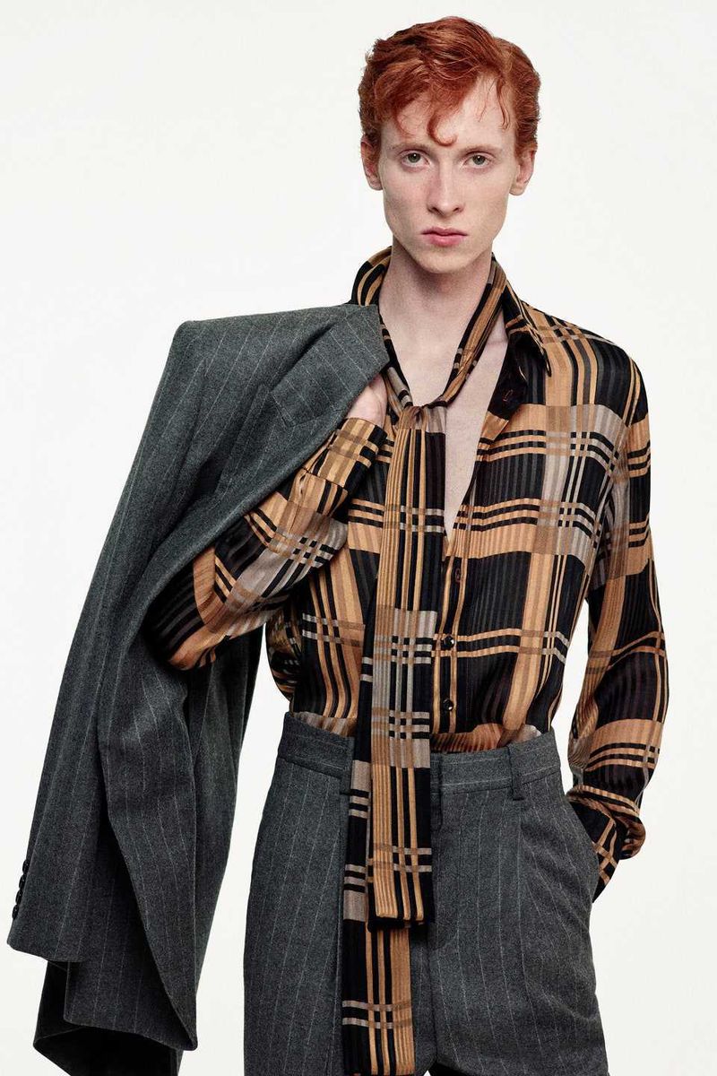 Male model with red hair poses in an unbuttoned patterned shirt and pinstripe pants, with a jacket draped over his shoulder.