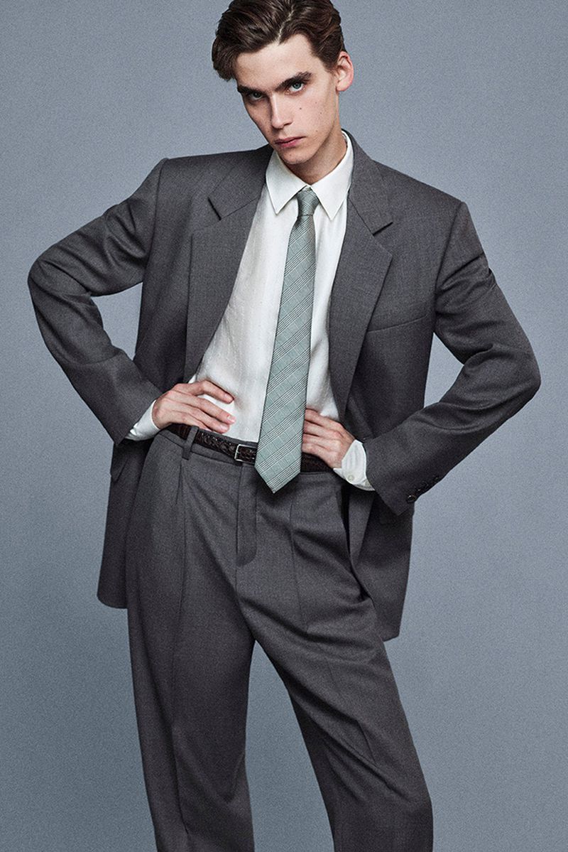 A young man poses in a grey suit with a white shirt and patterned tie, hands on hips, against a neutral background in a studio shot.