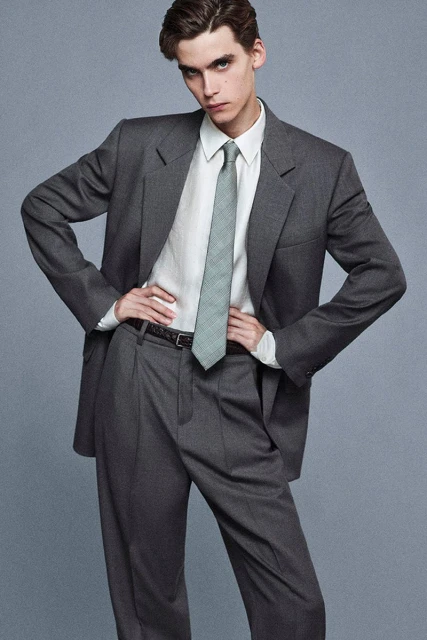 A young man poses in a grey suit with a white shirt and patterned tie, hands on hips, against a neutral background in a studio shot.