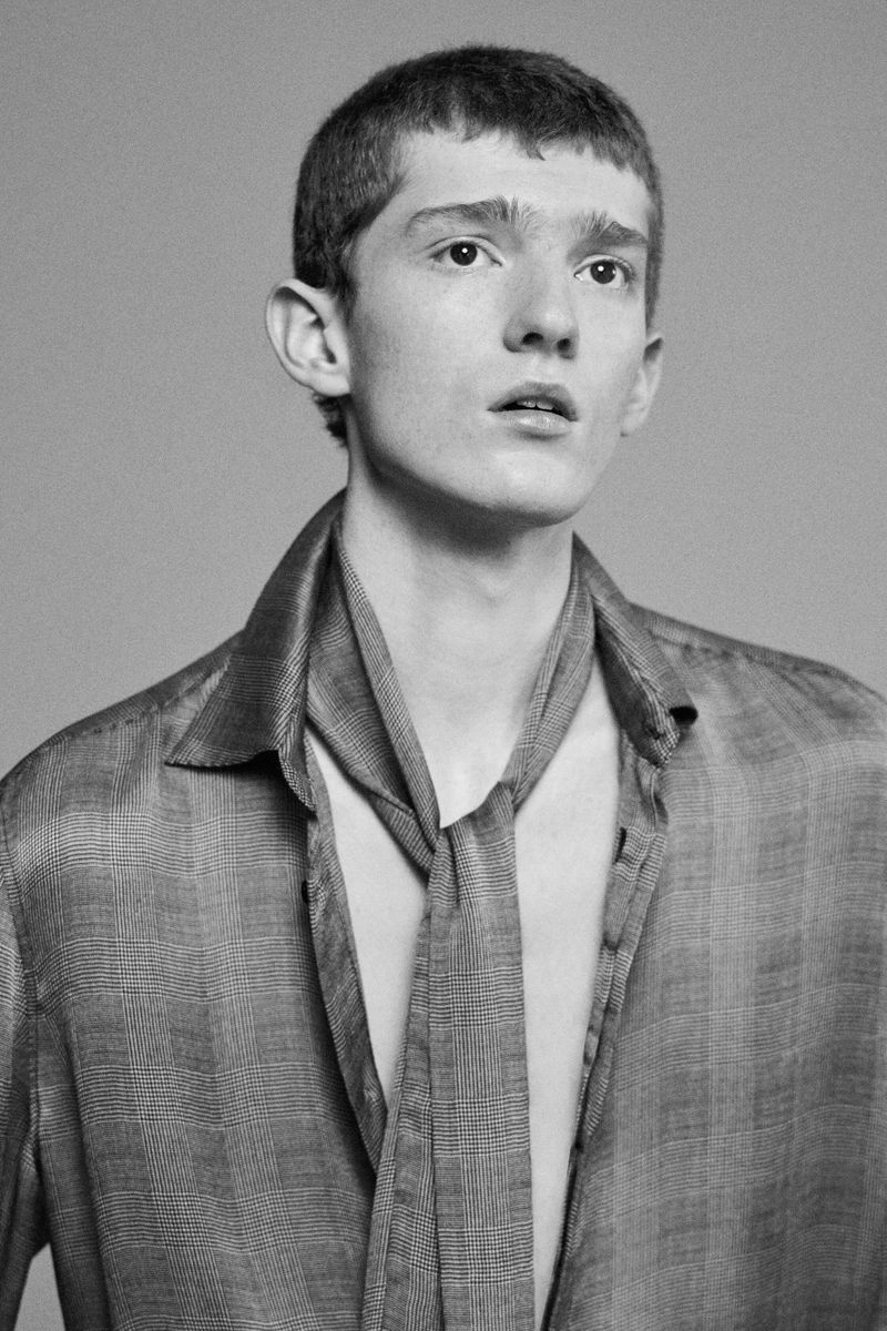 Black and white portrait of a young adult male with short hair wearing an unbuttoned shirt and a loosely tied necktie, looking upward.
