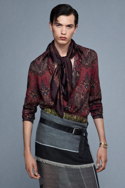 A young adult male poses in a paisley shirt and skirt ensemble, complemented by a black belt and brown scarf, against a neutral backdrop.